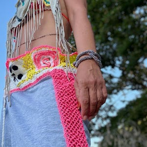 May include: A colorful crochet top and belt with a cat design, paired with gray sweatpants featuring a pink crochet stripe. The outfit includes a fringed scarf, sunglasses, and bracelets. The overall theme is bohemian.