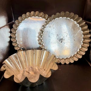 Puede incluir: Tres moldes para tartaletas vintage de color plateado. Dos son redondos con bordes festoneados, y uno es un molde acanalado con forma de flor. Los moldes son de metal y muestran signos de desgaste, lo que sugiere que son antiguos o muy usados.