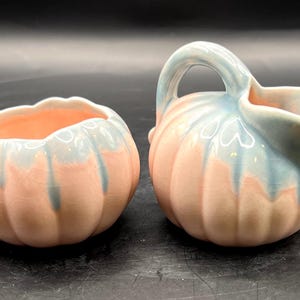 May include: A ceramic creamer and sugar bowl set. The pieces are pumpkin-shaped with a pink base and blue-green accents. The creamer has a handle and spout. The sugar bowl is open.