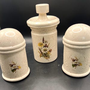 May include: A speckled ceramic kitchen set featuring a salt shaker, pepper shaker, and a jar with a lid. Each piece has a floral design with yellow and white flowers. The set is beige with brown speckles.