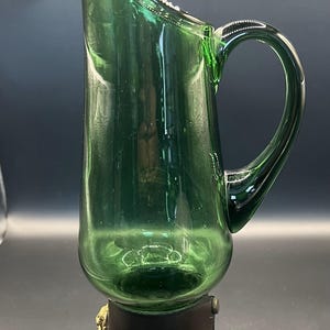 Könnte beinhalten: Eine grüne Glaskanne mit einer breiten Öffnung und einem Lederband um die Basis. Die Kanne hat ein einzigartiges Design mit einem gebogenen Ausguss und einem Griff.