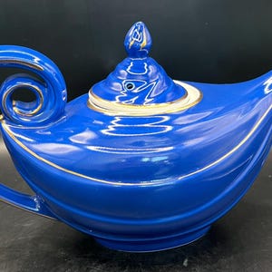 Puede incluir: Una tetera azul cobalto con un asa y un pico curvos, con adornos dorados en los bordes y la tapa. La tapa está rematada con un adorno dorado. La tetera tiene un acabado brillante y un diseño clásico y elegante.