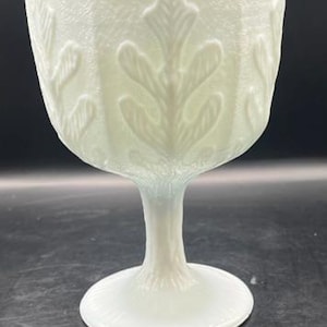 May include: A white, vintage-style goblet with a textured surface and leaf pattern. The goblet has a wide bowl, a slender stem, and a circular base. The leaf design is repeated around the bowl. The goblet is made of glass.