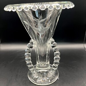 May include: Clear glass vase with a flared top and etched bird design. The vase has two beaded handles and a scalloped rim. The base is round and the vase is transparent, allowing light to pass through.