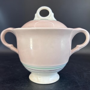 May include: A vintage pink sugar bowl with a matching lid. The bowl has two handles and a white base. The lid has a ring handle. The bowl is decorated with green and brown stripes near the base. A classic piece for serving sugar.