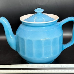 May include: A light blue ceramic teapot with a matching lid and handle. The teapot has a ribbed design on the body and a white interior rim. The spout curves gracefully, and the handle is a simple loop. A classic kitchenware item.