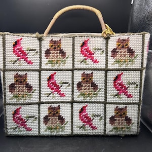May include: A white needlepoint handbag featuring a repeating pattern of red cardinals and brown owls. The bag has a woven handle and a gold-colored clasp. The design is framed by a dark green border.