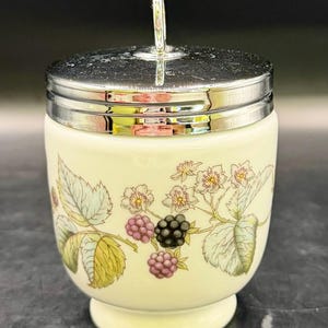 May include: A cream-colored ceramic egg coddler with a shiny silver-tone metal lid and a vertical ring handle. The body features a botanical illustration of green leaves, small white flowers with yellow centers, and clusters of purple and black blackberries.