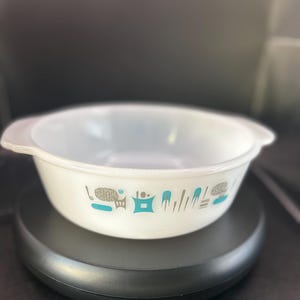 May include: White Pyrex bowl with handles, featuring a teal and gray pattern of stylized objects. The bowl is round and sits on a black surface. A vintage kitchenware item.