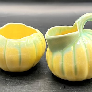 May include: A yellow and green ceramic creamer and sugar bowl set. The sugar bowl is round with a scalloped rim. The creamer has a handle and spout. Both pieces have a ribbed design and a yellow base with green accents.
