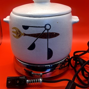 May include: White ceramic slow cooker with a brown and black design. The cooker has a lid and a cord with a plug. The base is silver metal.