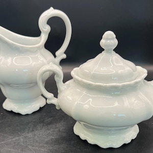 May include: A white porcelain creamer and covered sugar bowl set. The creamer has a curved spout and ornate handle. The sugar bowl has a lid with a decorative finial and two handles. Both pieces have a scalloped base.
