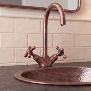 May include: A high-arc tap with a dark copper finish and two cross-style handles, paired with a matching round, hammered copper sink basin. The tap features a curved spout design and is mounted directly behind the circular metal basin.