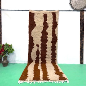 Op de afbeelding: Een rechthoekig tapijt met een bruin en beige streepmotief. Het tapijt heeft een getextureerd oppervlak en een franje langs de onderrand. Het tapijt wordt gepresenteerd tegen een witte muur en een groene vloer.