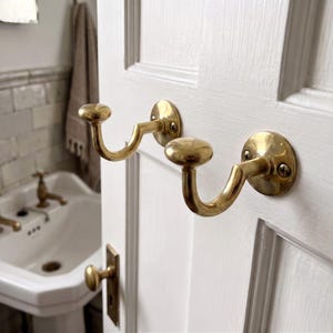 May include: Two polished brass wall hooks mounted on a white paneled door. Each hook features a curved arm with a rounded knob at the end and a circular base plate secured with two visible screws. The metal has a reflective, metallic gold finish.