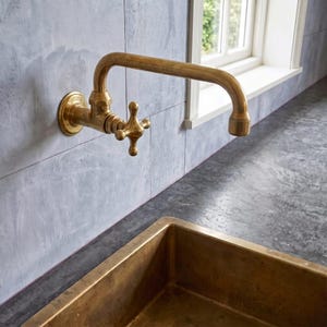 May include: A wall-mounted brass faucet with a cross-handle design and a curved spout, positioned above a rectangular brass sink basin. The faucet features a metallic finish and is attached to a light gray tiled wall surface in a kitchen area.