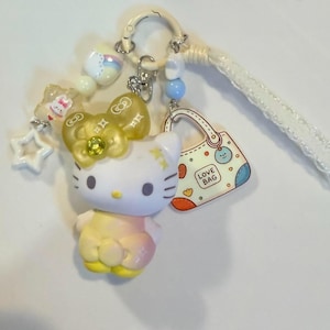May include: A keyring featuring a white cat figure with a yellow bow, a small handbag charm labelled LOVE BAG, and various star and bead accents. The cat figure has a yellow and pink gradient body, and the handbag charm is white with colourful patterns.