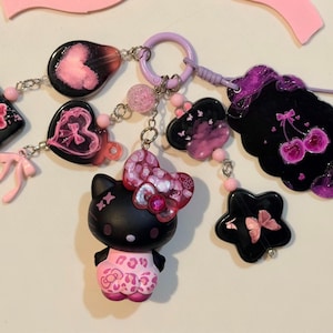 Peut inclure: Un porte-clés représentant une figurine de chat noir avec une tenue à motif léopard rose et un nœud rose pailleté. Le porte-clés comprend divers charms tels que des cœurs noirs et roses, une étoile avec un papillon et une plaque noire avec des cerises roses.