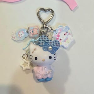 Peut inclure: Un porte-clés avec une figurine de chat blanc portant une robe rose clair à motif de visages de chat répétés et un nœud bleu à motifs. Le porte-clés comprend un anneau en forme de cœur et plusieurs petits breloques, dont une étoile blanche.