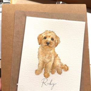 May include: A rectangular greeting card featuring a watercolor illustration of a golden-colored puppy sitting down. The puppy has fluffy fur, dark eyes, and a black nose. Below the puppy, the name Ruby is written in elegant black cursive script.