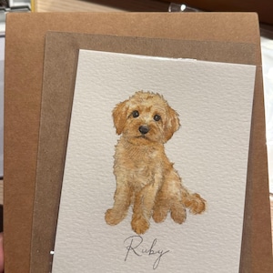 以下が含まれることがあります： ゴールデンカラーの子犬が座っている水彩画が描かれたグリーティングカード。子犬はふわふわした毛並み、暗い色の目、黒い鼻をしています。イラストの下には、エレガントな黒の筆記体でRubyという名前が書かれています。
