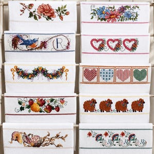 May include: A display of white embroidered towels with various designs, including flowers, birds, hearts, and sheep. The towels are arranged on a decorative rack, showcasing the detailed cross-stitch patterns and colorful threads. The towels are a home decor item.