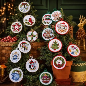 May include: A collection of embroidered Christmas ornaments, each in a round hoop. Designs include a snowman, angel, penguin, wreath, and a red cardinal. The ornaments are displayed on small Christmas trees, with a candle and decorative deer in the background.