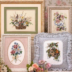 May include: A collection of framed cross-stitch art featuring floral designs. The artwork includes a basket of flowers, a bouquet, a teapot with flowers, and a birdhouse with floral accents. The frames are gold and white, with a decorative pillow in the foreground.