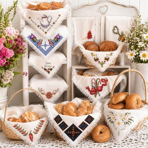 May include: A display of baked goods in wicker baskets and on shelves, lined with white embroidered cloths. The cloths feature various cross-stitch designs, including flowers, a teapot, and geometric patterns. Baskets are filled with bread, rolls, and muffins.