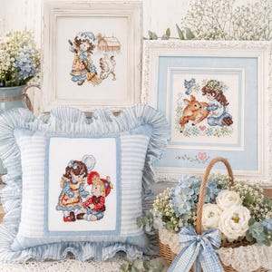 May include: A display of framed cross-stitch art and a decorative pillow. The art features children and animals in a vintage style. The pillow has a blue and white striped pattern with a ruffled edge. A basket of flowers with a blue bow completes the scene.