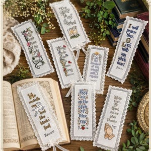 May include: A collection of handmade, embroidered bookmarks with white lace trim. Each bookmark features a different Bible verse and decorative elements like flowers, a lamb, and praying hands. The bookmarks are displayed on a wooden surface.