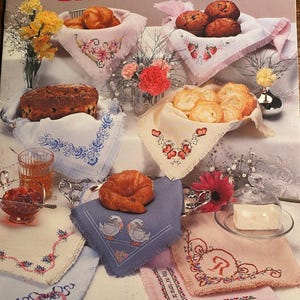 Peut inclure: Couverture de livre avec le texte "Sam Hawkins Designs Cross Stitch for BREAD CLOTHS". L'image montre plusieurs torchons brodés avec des motifs floraux et autres, et des paniers de pain et de pâtisseries.