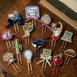 Puede incluir: Una colección de clips decorativos con diseños de punto de cruz. Los clips presentan varios motivos, incluyendo una casa, flores, un gato, un ordenador y una hamburguesa. Algunos clips tienen cintas adjuntas. El texto "Forget me not" es visible en un clip.
