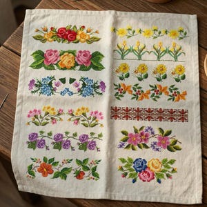 Può includere: Un tessuto color crema con vari disegni di bordi floreali ricamati. I disegni includono rose, margherite e altri fiori in tonalità di rosso, rosa, giallo, blu e arancione. Il ricamo è dettagliato e colorato.