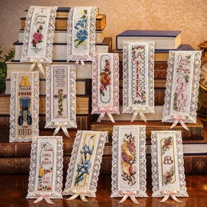 May include: A collection of embroidered bookmarks with lace trim and pink ribbons. The bookmarks feature floral designs, text like "Someone Special," "A Friend," and personalized messages. They are displayed on a stack of books.