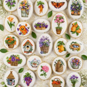 May include: A collection of oval-shaped embroidered fabric ornaments featuring various botanical, floral, and garden-themed designs including flowers, fruits, vegetables, butterflies, birdhouses, and a teapot, all framed with a gold twisted cord.