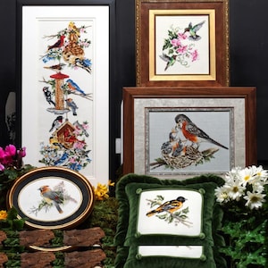 May include: A collection of framed cross-stitch art featuring birds. The artwork includes a tall piece with a bird feeder, a framed piece with hummingbirds, and a framed piece with a robin feeding its young. A green velvet pillow with a bird design is also present.