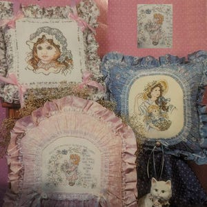 Peut inclure: Plusieurs coussins brodés au point de croix avec des motifs floraux et des portraits. Les coussins présentent des images de femmes et d'enfants avec des bonnets et des chapeaux, entourés de motifs floraux. Les coussins ont des bords à volants et sont dans des tons de rose et de bleu.
