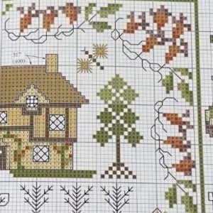 May include: Close-up of a cross-stitch pattern featuring a house, trees, and floral elements. The house has a brown roof and tan walls with windows. The trees are green and brown. The floral elements include leaves and acorns in shades of brown and green.