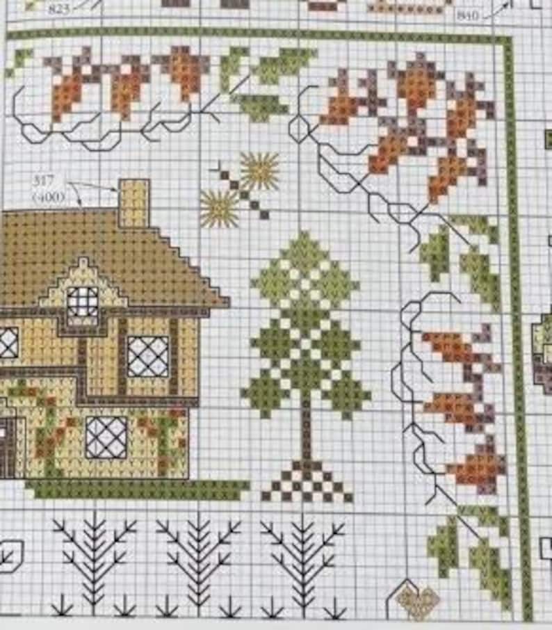 May include: Close-up of a cross-stitch pattern featuring a house, trees, and floral elements. The house has a brown roof and tan walls with windows. The trees are green and brown. The floral elements include leaves and acorns in shades of brown and green.
