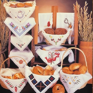 May include: A display of baked goods in baskets and on shelves, lined with white cloths featuring cross-stitch embroidery. The cloths showcase various designs, including a teapot, butterflies, and holiday motifs. Baskets contain muffins, rolls, and bagels.