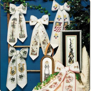 May include: A collection of decorative, embroidered ribbon bows and framed cross-stitch art. The bows feature various designs, including crosses, angels, shamrocks, and patriotic stars. A framed cross-stitch of a witch is also present.