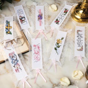 May include: A selection of embroidered bookmarks, each with a unique design. The white bookmarks feature floral and nature motifs, some with text. They are finished with pink ribbons and lace edging, ideal for marking pages.