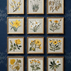 Puede incluir: Una colección de doce obras de arte florales en punto de cruz enmarcadas. Cada pieza presenta una flor diferente en tonos de amarillo, blanco y verde, sobre un fondo claro. Los marcos son dorados y las obras de arte se exhiben en una pared azul oscuro estampada.