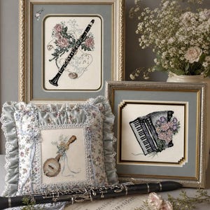 Op de afbeelding: Een verzameling ingelijste kruissteekkunst met muziekinstrumenten en bloemstukken. Bevat een klarinet, banjo en accordeon. Er is ook een decoratief kussen met een banjomotief. Het kleurenpalet omvat zachte blauwtinten, roze en crèmes.