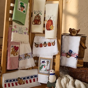 Peut inclure: Un présentoir en bois présente des serviettes décoratives brodées. Les motifs incluent un lutin, des citrouilles, des ours et des thèmes patriotiques. Les serviettes sont blanches, rouges, vertes et bleues, avec des bords effilochés. Certaines serviettes ont du texte.