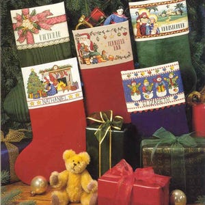 May include: Several Christmas stockings in red, green, and purple, each with embroidered festive designs and names. The stockings feature images of Santa Claus, snowmen, and a nativity scene. A teddy bear and wrapped gifts are also visible in the image.