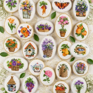 Könnte beinhalten: Eine Sammlung von ovalen Stickereien mit verschiedenen Blumen- und Naturmotiven. Designs umfassen Blumen in Körben, einen Schmetterling, ein Vogelhaus, Früchte und eine Teekanne, alle mit goldfarbenem Seilbesatz.