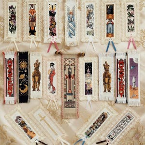 May include: Embroidered bookmarks featuring diverse designs such as flowers, a fox, butterflies, an angel, and a rocket. Each bookmark is framed with lace and has a ribbon attached. One bookmark displays the word "FABLES".