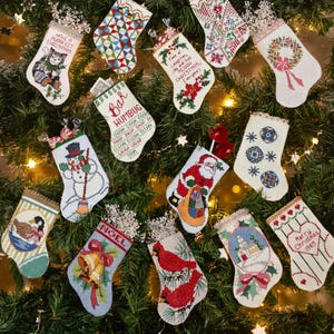 May include: A collection of miniature Christmas stockings, each with unique embroidered designs. The stockings feature various festive motifs, including a cat, snowman, Santa Claus, and cardinals. Some stockings have text like "NOEL" and "Merry Christmas 1989".